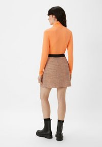 Langarmige oranje top met een hoge kraag, gecombineerd met een rokje met een houndstooth-patroon. Zwarte tailleband en kniehoge zwarte laarzen.