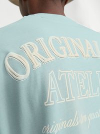 T-shirt bleu clair avec un texte en relief blanc « ORIGINAL ATELIER » au dos, avec une texture de tissu lisse.