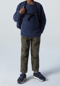 Granatowy sweter w prążki, oliwkowe spodnie kordowe i czarne trampki z zielonymi akcentami. Plecak z widocznymi paskami.