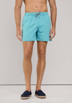 Shorts de bain bleu clair avec une taille élastique et un cordon de serrage, se distinguant par un tissu texturé. Associés à des chaussures sombres à enfiler.