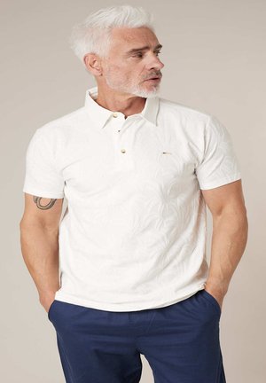 Man met grijs haar en baard, gekleed in een wit poloshirt met patroon en marineblauwe broek, handen in de zakken, kijkt opzij.
