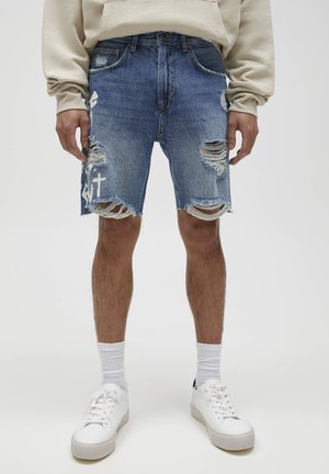 Shorts en denim d'un bleu délavé, avec des déchirures usées et des bords effilochés. Associés à des baskets blanches et des chaussettes blanches côtelées.