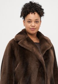 Brązowy płaszcz z futra sztucznego z szerokim kołnierzem, o miękkiej i gładkiej fakturze. Płaszcz ma luźny krój i jest zaprojektowany w średniej długości.