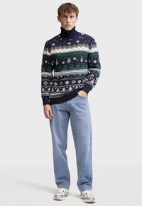 Marineblauer Strickrollkragenpullover mit gemusterten Designs in Weiß, Grün und Grau. Kombiniert mit hellblauen Jeans und sportlichen Sneakers.