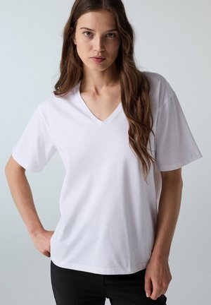 Frau mit langen braunen Haaren, die ein schlichtes weißes V-Ausschnitt-T-Shirt und schwarze Hosen trägt, steht vor einem hellgrauen Hintergrund.