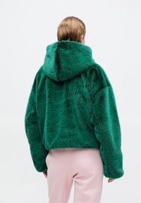 Grüne Fake-Fur-Jacke mit Kapuze, cropped Design und strukturierter Oberfläche, kombiniert mit hellrosa Hosen. Die Rückansicht zeigt eine voluminöse Passform.