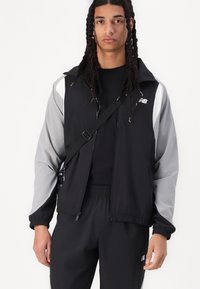 Black and gray windbreaker jacket with a zip front, elastic cuffs, and logo detail. Smooth texture, sporty design, worn with matching pants.