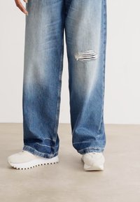 Jeans in denim azzurro chiaro, vissuti e con una vestibilità ampia, con orli sfrangiati visibili sulle ginocchia, abbinati a sneakers sportive bianche.