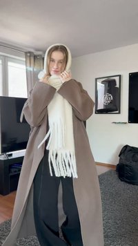 Brauner, übergroßer Mantel mit weiten Ärmeln, kombiniert mit einem weißen, fransigen Schal. Der Schal hat ein strukturiertes Muster. Schwarze Hosen sind darunter sichtbar.