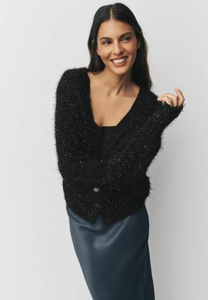 Sort fuzzy cardigan med dyb V-hals, knaplukning og lange ærmer, parret med en glat, blå satin-nederdel. Teksturert og hyggelig udseende.