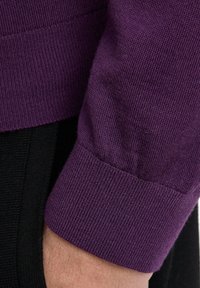 Cuff di un maglione viola lavorato a maglia con texture a coste e design aderente, in contrasto con uno sfondo nero.