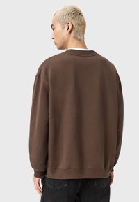 Sudadera marrón de corte holgado, con puños acanalados y cuello redondo. El material parece suave y ligeramente texturizado. Combinada con vaqueros oscuros.