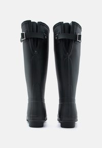 Μαύρες γαλότσες από καουτσούκ μέχρι το γόνατο με ρυθμιζόμενους ιμάντες και ασημένιες αγκράφες, εμφανιζόμενες από πίσω σε λευκό φόντο.