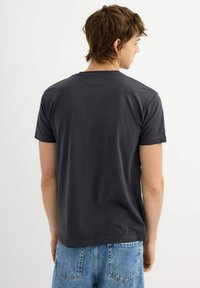 Camiseta de algodón gris oscuro con cuello redondo clásico, mangas cortas y un corte relajado, mostrada desde una perspectiva trasera.