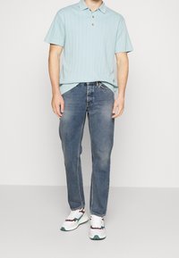 Polo azzurro chiaro a maniche corte con colletto a tre bottoni, abbinato a jeans blu medio e sneakers bianche con accenti multicolori.