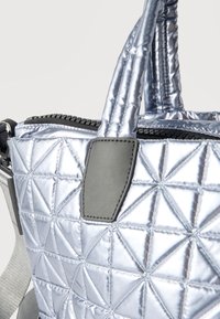 Καπιτονέ μεταλλική ασημί τσάντα tote με επενδυμένες λαβές, μαύρο φερμουάρ και γκρι δερμάτινες λεπτομέρειες σε λευκό φόντο.