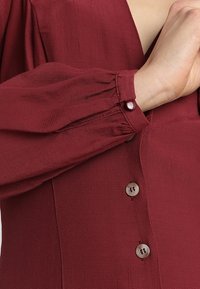 Burgundin yläosa, jossa on pitkät, pleaatit hihat ja nappi mansetit. Ominaisuuksiin kuuluu teksturoitu kangas ja kaksi nappia etulistan varrella.