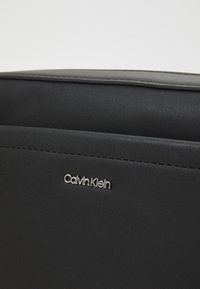 Juodas sintetinio odos krepšys su lygia tekstūra, su subtiliu sidabriniu "Calvin Klein" logotipu ir minimalizmo dizaino detalėmis.