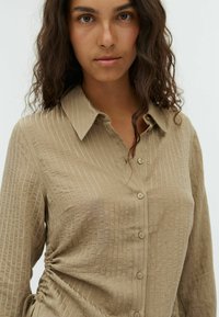 Donna con lunghi capelli ricci che indossa una camicia beige con bottoni, caratterizzata da righe verticali e un dettaglio arricciato sul lato, rivolta in avanti con un'espressione neutra.