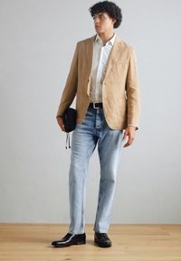 Blazer en lin beige sur une chemise blanche, associé à un jean bleu clair et des chaussures en cuir noires. Tient un sac noir, debout sur un sol en bois.