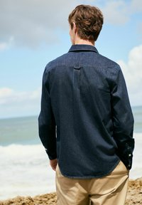 Donkerdenim shirt met lange mouwen, knoopsluiting bij de mouwen en een licht gebogen zoom. Draagt bij lichte broek, met een kustachtergrond.