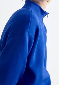 Modrá fleecová mikina s vysokým límcem a polovičním zipem, s klesajícími rameny a měkkou, texturovanou tkaninou.