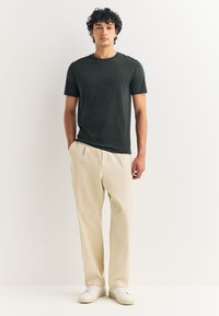 Temno zelena kratka majica z okrolim izrezom, kombinirana z lahkimi bež hlačami in belimi supergami, stoječa na nevtralnem ozadju.