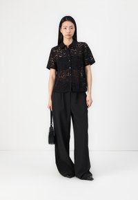 Chemise en dentelle noire à manches courtes avec un devant à boutons, portée avec un pantalon large noir. Les accessoires comprennent un petit sac à main noir.