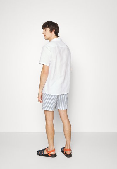 Camicia bianca a maniche corte con una vestibilità rilassata, abbinata a pantaloni corti a righe blu e bianche e sandali neri con dettagli arancioni.
