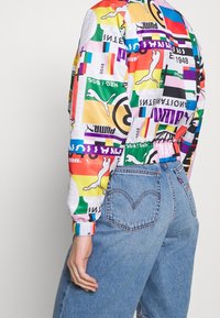 Kurtka Puma z wielokolorowego poliestru z żywymi wzorami, elastycznymi mankietami i ściągaczem w talii, w zestawieniu z jasnoniebieskimi dżinsami.