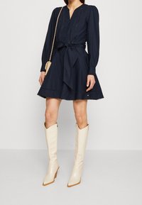 Abito navy con maniche lunghe, chiusura a bottoni davanti e cintura in vita. Presenta una gonna svasata e dettagli in pizzo. Abbinato a stivali bianchi alti e lucidi.