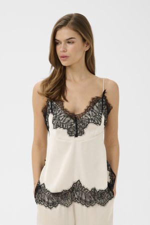 Kvinde iført hvid satin camisole med sort blonderkant, stående med hænderne i lommerne, kiggende let mod venstre mod en ensfarvet baggrund.