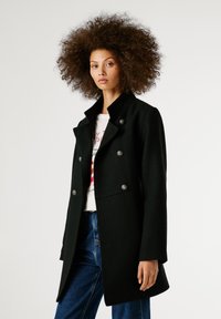 Melna divrindota mētelis ar augstu apkakli, struktūras vilnas audumu, sudraba krāsas pogām un taisnu siluetu.