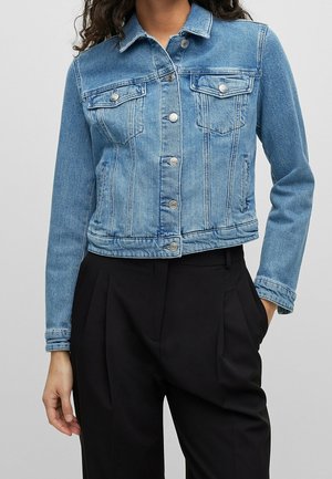 Donna con giacca corta in denim blu con bottoni d'argento e pantaloni neri a vita alta, mano in tasca, su sfondo grigio.