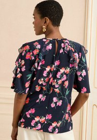 Blusa blu navy con motivo floreale e maniche a volant, decorata con fiori rosa e arancioni, e un piccolo dettaglio a chiave sul retro del collo.