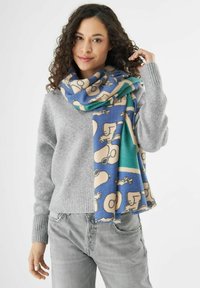 Écharpe à motifs en bleu, beige et vert, avec des designs de style cartoon. Fabriquée en matériau doux, elle tombe librement sur un pull gris.