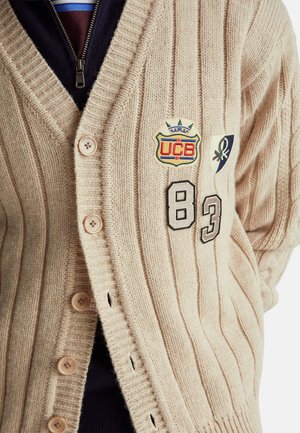 Beige gestrickte Strickjacke mit geripptem Muster, vier Knöpfen, bestickten Aufnähern und den schwarzen Zahlen 83 und 8.