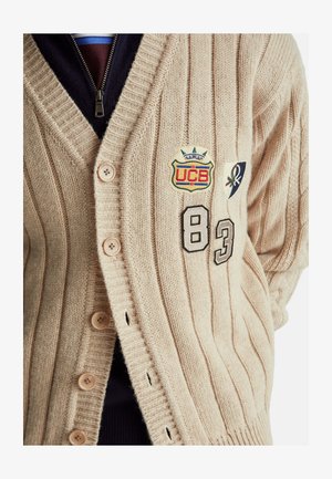 Beige gestrickte Strickjacke mit geripptem Muster, vier Knöpfen, bestickten Aufnähern und den schwarzen Zahlen 83 und 8.