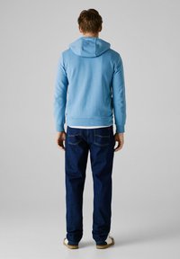 Ljust blå huvtröja med en slät yta, bärs över en vit skjorta. Matchad med mörkblå jeans och beige skor.