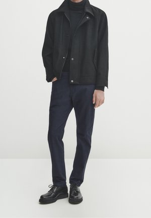 Mann trägt schwarze Jacke, schwarzen Rollkragenpullover, marineblaue Hose und schwarze Schnürschuhe, steht mit einer Hand in der Tasche.