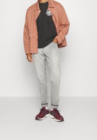 Osoba w rudawym płaszczu, czarnym t-shircie z grafiką, jasnoszarych dżinsach z podwiniętymi nogawkami, białych skarpetkach i bordowych sneakersach na jednolitym tle.