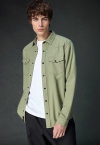 Olive green button-up shirt with two chest pockets, black snap buttons, and a classic collar. Worn over a white T-shirt. Black trousers.