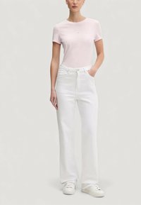 Světle růžové tričko s krátkým rukávem a bílé džíny s vysokým pasem a rovnými nohavicemi, opatřené knoflíkem a zipem vpředu. Bílé tenisky.