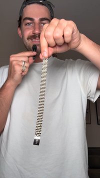 Metallarmbånd til klokke med vekslende gull- og sølvlenker, presentert i en hånd; har clasp og en logotagg på enden.