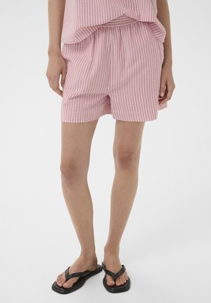 Žena nosiaca ružové a biele vertikálne pruhované šortky a zodpovedajúci top, v kombinácii s čiernymi žabkami so štvorcovou špičkou, stojaca na bielom pozadí.