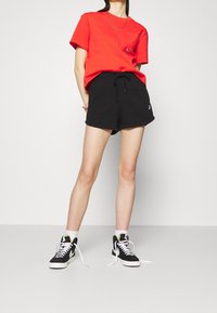 Red cotton t-shirt with a pocket and logo, paired with black sweat shorts. Black high-top sneakers with white soles and laces.