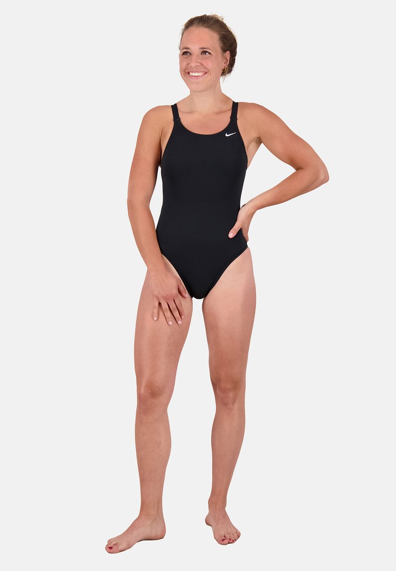 Maillot de bain une pièce noir avec des fines bretelles, design ajusté, texture lisse et un petit logo blanc sur la poitrine. Modèle debout pieds nus.