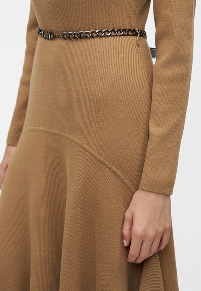 Gros plan d’une personne portant une robe en maille côtelée couleur camel, avec une ceinture en chaîne de métal foncé et des coutures incurvées.