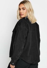 Mustat farkkutakki, jossa on hieman yli kokoa oleva istuvuus, näkyvät saumat ja napit hihansuissa. Suuri tekstuuri ja minimalistinen muotoilu.