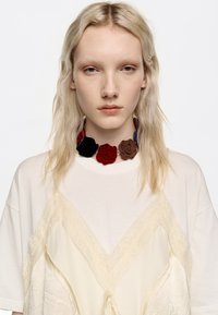 Przesadna biała koszulka z przezroczystymi kremowymi koronkowymi akcentami i związanym detalem. Dekoracyjny naszyjnik z kwiatami w kolorach czerwonym, czarnym, brązowym i niebieskim.